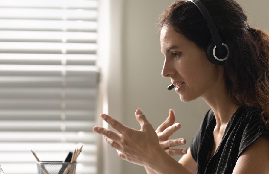 Person wearing a headset speaking and gesturing while using a computer at a desk.