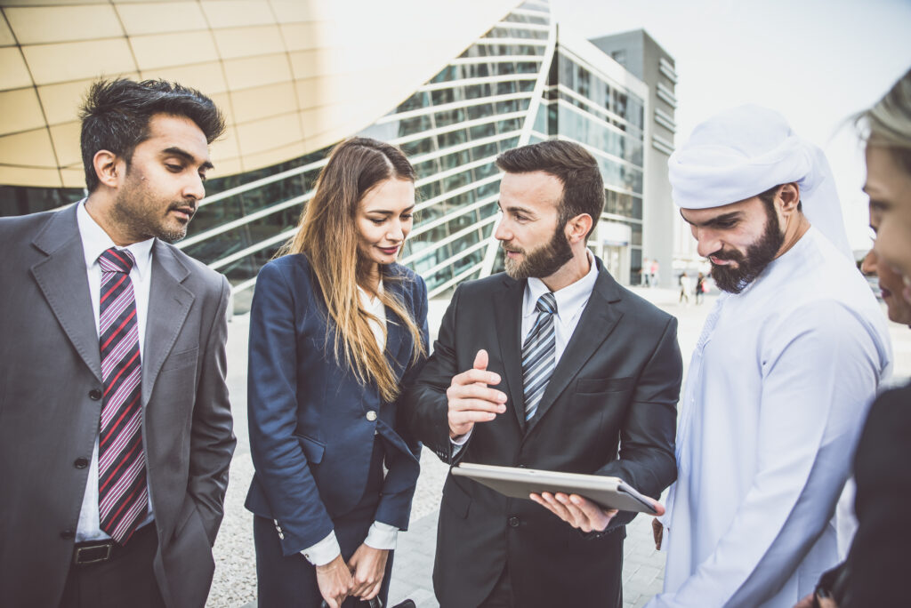 Four people stand outside an office building reviewing a tablet during a conversation.