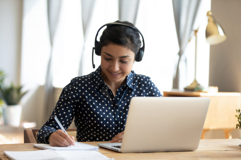 Person wearing headphones writes notes while using a laptop at a desk indoors.