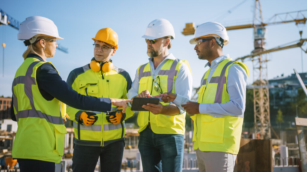 Four people in safety helmets and high-visibility clothing review a tablet at a construction site.