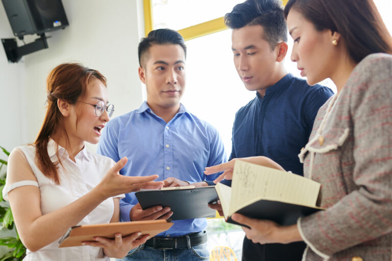 Four people stand together reviewing a tablet and notebook while discussing work indoors.