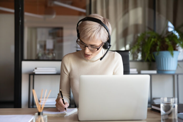 Person wearing a headset writes notes while using a laptop at a desk indoors.