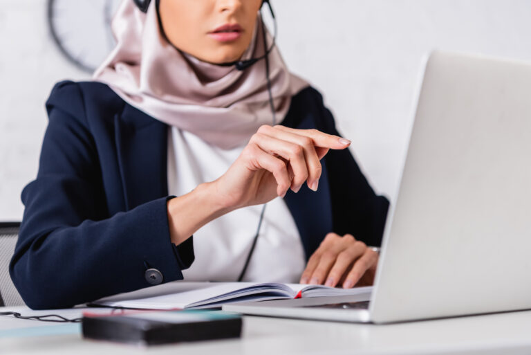Person wearing a headset uses a laptop and points while reviewing notes at a desk.