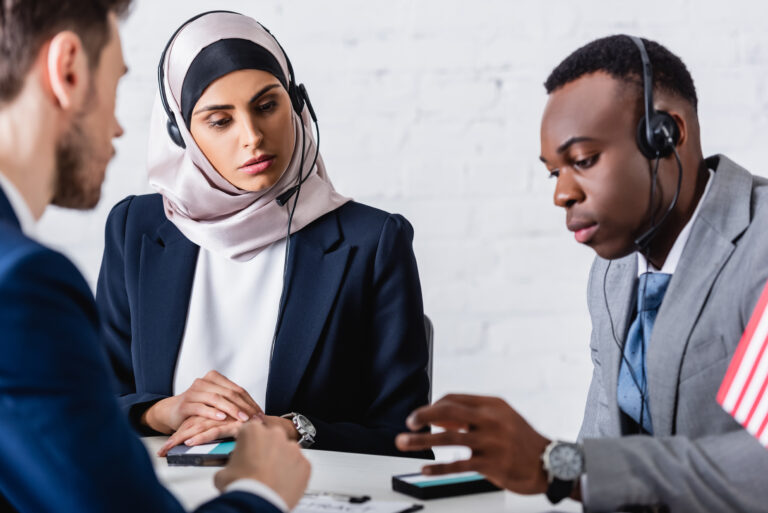 Three people wearing headsets sit at a desk, reviewing devices and discussing work together.