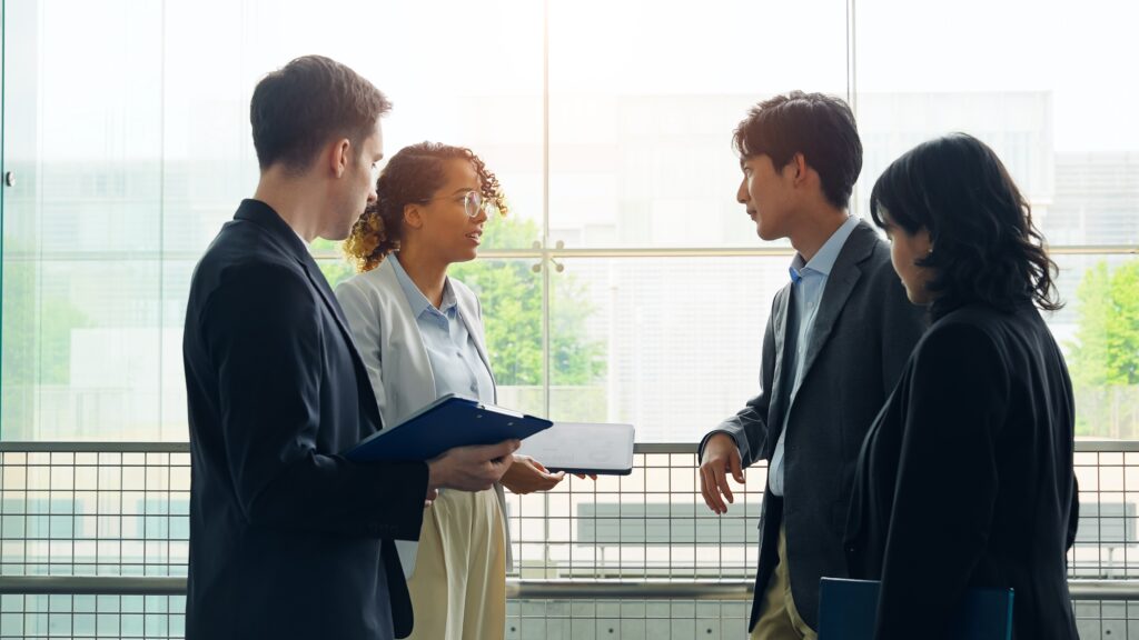 Four people stand together indoors discussing work, with one holding a folder during conversation.