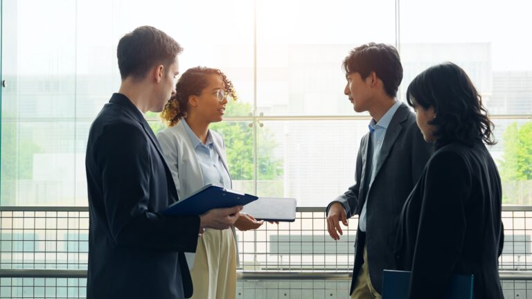 Four people stand together indoors discussing work, with one holding a folder during conversation.