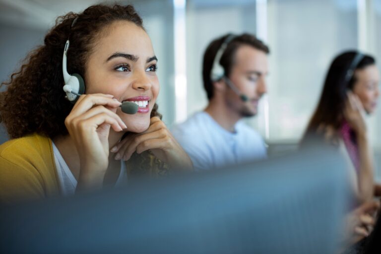 People wearing headsets sit at computers, working and speaking in an office environment.